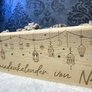 Op de afbeelding: Een houten doos met een Ramadan-kalenderontwerp. De doos is versierd met sterren, halve manen en lantaarns. De tekst "Ramadankalender von" is in de doos gegraveerd.