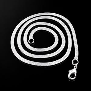 Op de afbeelding: Een zilverkleurige visgraatketting opgerold in een cirkelvormig patroon. De platte, flexibele schakels creëren een glad, reflecterend oppervlak. Aan het ene uiteinde zit een kleine ronde ring en aan het andere uiteinde een karabijnsluiting.
