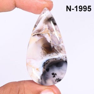 Puede incluir: Una piedra preciosa en forma de lágrima con una mezcla de patrones blancos, beige, negros y grises. La piedra se sostiene entre los dedos y tiene el texto "N-1995" en la esquina superior derecha. La superficie de la piedra tiene un aspecto veteado.