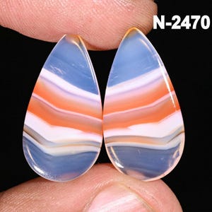May include: Two teardrop-shaped gemstones with layered bands of blue, orange, white, and grey. The stones are held between fingers against a black backdrop. The text "N-2470" is visible in the upper right corner.