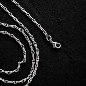 May include: A silver-toned metal chain necklace featuring elongated, cylindrical links connected by small circular rings. The chain includes a secure lobster claw clasp mechanism at one end, allowing for easy fastening and adjustment of the jewelry piece.