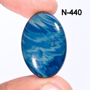 May include: An oval-shaped gemstone with a swirling pattern of blue and white. The stone is held between two fingers and has the text "N-440" in the upper right corner. The polished surface reflects light, highlighting the natural patterns.