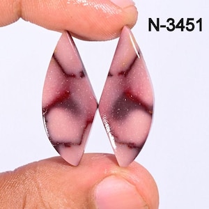 Puede incluir: Dos gemas en forma de lágrima con un tono rojizo-rosado y vetas rojas oscuras. Las piedras se sostienen entre los dedos sobre un fondo blanco. El texto "N-3451" es visible en la esquina superior derecha.