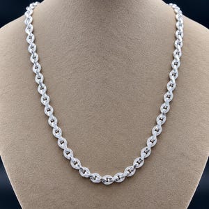Peut inclure: Un collier chaîne de couleur argentée composé d'une série de maillons ovales entrelacés. Chaque maillon individuel présente un motif texturé à hachures croisées sur sa surface, créant un design répétitif sur toute la longueur de la chaîne.