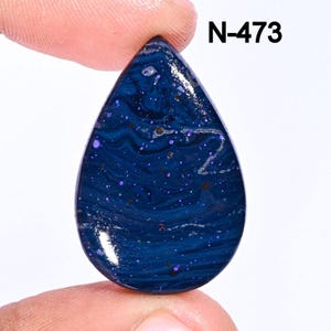 May include: A teardrop-shaped cabochon gemstone with a deep blue base and swirling patterns of white and purple. The stone has a speckled appearance with small, scattered purple flecks. The image includes the text "N-473".