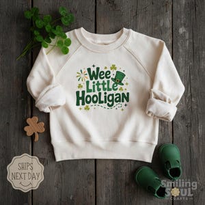 May include: Cream-colored sweatshirt with the text "Wee Little Hooligan" in green, featuring a leprechaun hat and shamrocks. The sweatshirt is paired with green baby shoes and a wooden shamrock cutout, perfect for St. Patrick's Day.