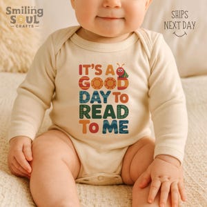 May include: A beige long-sleeved baby bodysuit with the text "IT'S A GOOD DAY TO READ TO ME" in colorful letters. The design includes a cartoon caterpillar and sun. The Smiling Soul Crafts logo is in the upper left corner.