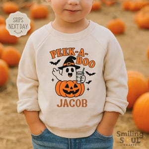 May include: A beige Halloween-themed sweatshirt with the text "PEEK-A-BOO" in orange and a ghost character wearing a witch hat and holding a drink. The ghost is sitting on an orange pumpkin. The name "JACOB" is printed below the pumpkin.