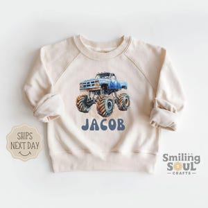 May include: Cream-colored sweatshirt featuring a blue monster truck graphic with the name "JACOB" in blue. The truck has large tires and is set against a watercolor style background. The sweatshirt has long sleeves and a crew neck.