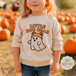 May include: A cream-colored sweatshirt with the text "LITTLE BOO BEAR" in orange lettering. The design features a ghost wearing an orange bear hat. The sweatshirt is worn by a child in a pumpkin patch.