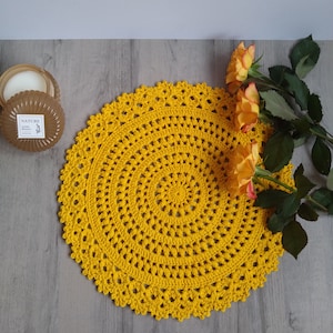 Può includere: Un centrino all'uncinetto giallo con un design circolare. Il centrino è su una superficie grigia con fiori gialli.