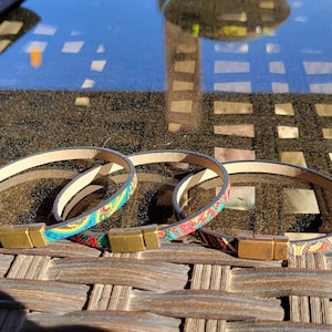 May include: Three leather bracelets with colorful patterned designs. Each bracelet has a gold-colored rectangular clasp. The bracelets are displayed on a brown woven surface, with a remote control and a reflective glass table in the background.