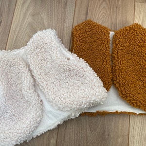 Könnte beinhalten: Zwei Kinderwesten. Eine ist cremefarben, die andere braun. Beide Westen bestehen aus einem flauschigen, strukturierten Material. Die Westen sind vorne offen und scheinen mit weißem Stoff gefüttert zu sein. Die Westen liegen auf einer Holzoberfläche.