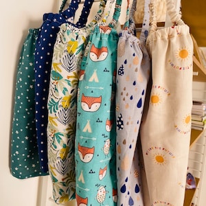 Puede incluir: Seis bolsas de la compra reutilizables colgadas de un estante blanco. Las bolsas son de tela y tienen diferentes patrones, incluyendo estrellas, animales y un mensaje que dice "Somos el futuro".