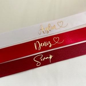 May include: Three satin ribbons with gold lettering. The top ribbon is white with the name "Aylin" and a heart. The middle ribbon is red with the name "Deniz" and a heart. The bottom ribbon is burgundy with the name "Serap".