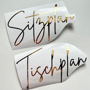 Puede incluir: Dos pegatinas blancas con letras doradas. La pegatina superior dice "Sitzplan" y la pegatina inferior dice "Tischplan".