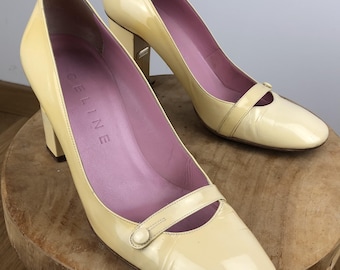 May include: A pair of pale yellow patent leather pumps with a block heel and a decorative strap across the vamp featuring a small button detail. The interior lining is a soft lavender color, and the Celine brand name is embossed on the insole.
