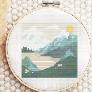 May include: Cross-stitch embroidery hoop with a landscape scene. The design features a blue and green mountain range, a body of water, and a yellow sun in the sky.