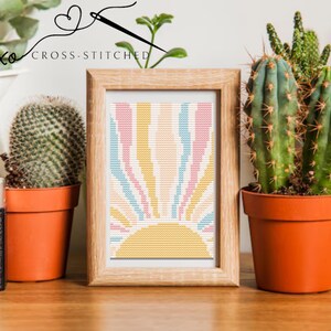 May include: A framed cross-stitch pattern of a colorful sun with a yellow center and pink, blue, and yellow stripes. The frame is made of wood and has a light brown finish. The image is surrounded by potted cacti.