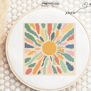 May include: A cross-stitch pattern of a colorful sunburst design with a yellow sun in the center. The pattern is framed in a white embroidery hoop.