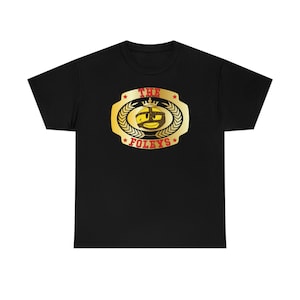 May include: Black t-shirt with a gold championship belt design featuring a cartoon face with a crown and the text "THE FOLEYS".