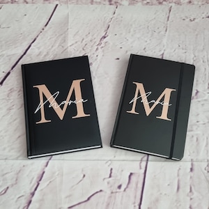 Könnte beinhalten: Zwei schwarze Notizbücher mit einem roségoldenen Buchstaben "M" und dem Namen "Maria" in weißer Schrift. Die Notizbücher haben ein schlichtes, elegantes Design, ideal für den persönlichen Gebrauch oder als Geschenk.