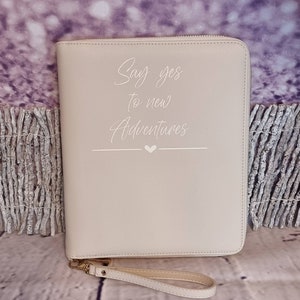 Könnte beinhalten: Eine beige Reißverschlusstasche mit einem weißen Riemen. Die Tasche trägt den Schriftzug "Say yes to new Adventures" in weißer Kursivschrift.