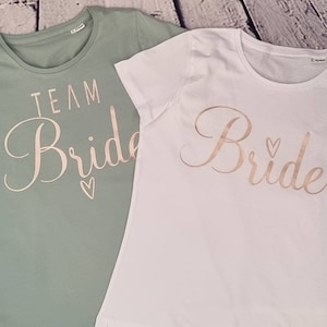 Könnte beinhalten: Zwei T-Shirts, eines grün und eines weiß, mit den Worten "Team Bride" und "Bride" in goldener Schrift. Das grüne Shirt hat ein kleines Herzmotiv.