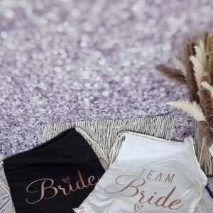 Op de afbeelding: Twee witte trekkoordtassen met zwarte handvatten. Een tas is zwart met het woord "Bride" in roségouden letters. De andere tas is wit met de woorden "Team Bride" in roségouden letters.