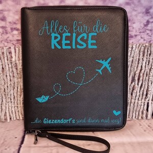 Könnte beinhalten: Schwarze Reise-Clutch mit Reißverschluss und einem blauen Flugzeug-, Herz- und Papierboot-Design. Der Text "Alles für die REISE...die Giezendort's sind dann mal weg!" ist auf der Vorderseite aufgedruckt.