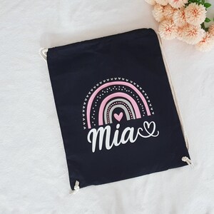 Könnte beinhalten: Eine schwarze Kordeltasche mit einem rosa und grauen Regenbogenmuster und dem Namen "Mia" in Weiß mit einem Herz.