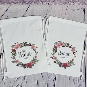 Op de afbeelding: Twee witte trekkoordtassen met bloemkransen en de tekst "Team Braut Nadia" en "Braut Jana".