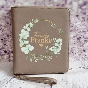 Könnte beinhalten: Ein brauner Lederimitat-Ordner mit Reißverschluss, einem goldenen Blumengirlanden-Design und dem Text "Familie Franke" in goldenen Buchstaben.