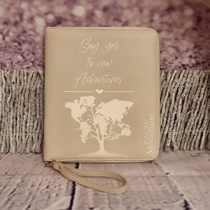 Könnte beinhalten: Beigefarbenes Leder-Reisetagebuch mit Reißverschluss und Trageriemen. Der Umschlag zeigt eine gravierte Weltkarte mit einem Baum und dem Text "Say yes to new Adventures". Der Text "Adventure Moosmann" ist ebenfalls auf dem Umschlag graviert.