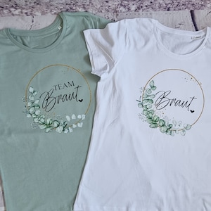 Könnte beinhalten: Zwei T-Shirts mit einem goldenen Kreis und einem Design aus grünen Blättern. Ein Shirt ist hellgrün mit dem Text "Team Braut" und das andere ist weiß mit dem Text "Braut".