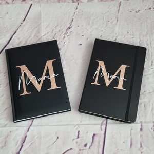 Könnte beinhalten: Zwei schwarze Notizbücher mit goldener Beschriftung. Die Notizbücher tragen den Namen "Maria" in Kursivschrift mit einem großen goldenen "M" davor.