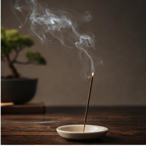 May include: A burning incense stick in a small, cream-coloured ceramic dish, releasing trails of white smoke. A bonsai tree in a dark pot is in the background, set on a wooden surface, creating a tranquil scene.