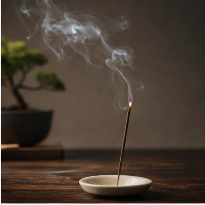 May include: A burning incense stick in a small, cream-coloured ceramic dish, releasing trails of white smoke. A bonsai tree in a dark pot is in the background, set on a wooden surface, creating a tranquil scene.