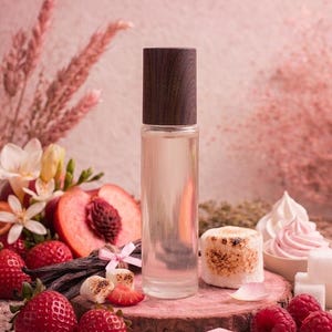 May include: A clear glass perfume bottle with a dark wood cap sits on a wooden surface, surrounded by fresh strawberries, raspberries, a sliced peach, vanilla beans, and toasted marshmallows. The background is a soft pink.