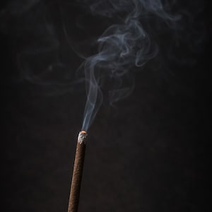 May include: A burning incense stick with smoke trails against a dark backdrop. The incense is brown with a white ash tip, and the smoke is light grey. The image suggests a tranquil atmosphere.