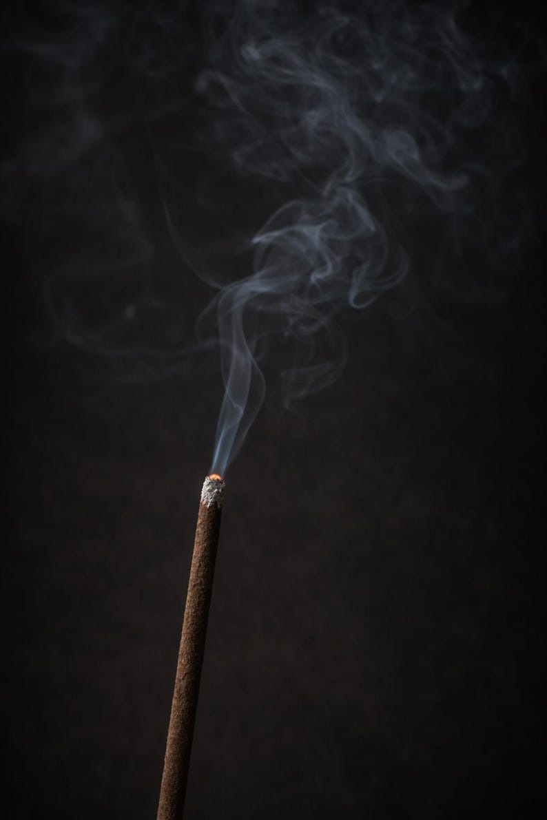 May include: A burning incense stick with smoke trails against a dark backdrop. The incense is brown with a white ash tip, and the smoke is light grey. The image suggests a tranquil atmosphere.