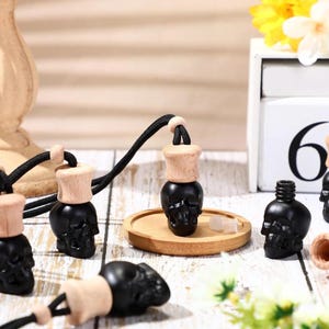 May include: Black skull-shaped car air fresheners with wooden tops and black cords. The air fresheners are displayed on a wooden tray and a white surface. The image also includes a wooden decorative piece and a calendar.