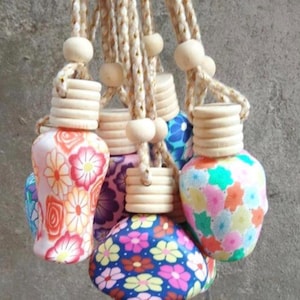 May include: A selection of hanging car air fresheners with floral patterns in various colours. Each freshener has a wooden cap and is suspended by a beige rope with a wooden bead. The designs include pink, blue, and yellow flowers.