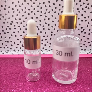 May include: Two clear glass dropper bottles with white labels. One bottle is labeled "10 ml." and the other is labeled "30 ml." Both bottles have gold-colored collars and white droppers. The bottles are on a pink glitter surface.