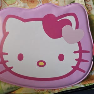 May include: A pink Hello Kitty-themed case with a white face, pink bow, and heart accents. The case has a rounded rectangular shape and includes a set of keys attached to a pink cord. The case is on a patterned surface.