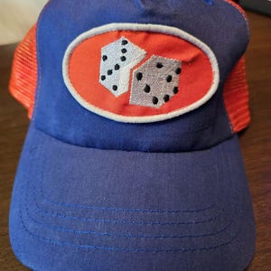 May include: A blue trucker hat featuring a red oval patch on the front panel. The patch displays an embroidered graphic of two light gray dice with black pips. The hat has a solid blue front, a curved brim, and bright orange mesh side panels.