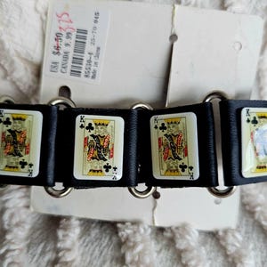May include: Black bracelet featuring playing card designs of the King of Clubs. The bracelet is made of black material with silver-tone metal rings connecting the playing card segments. The playing cards have a white background with a colorful design.