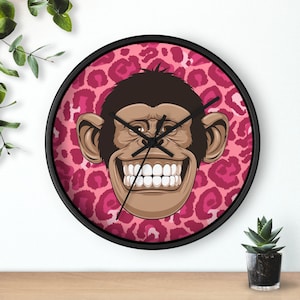 May include: A pink and black round wall clock with a cartoon monkey face with a big smile and white teeth. The monkey face is on a pink background with a leopard print pattern.