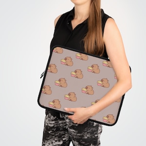 Könnte beinhalten: Eine braune Laptoptasche mit einem sich wiederholenden Muster aus Comic-Capybaras, die Popcorn essen. Die Capybaras sind braun mit weißen Akzenten und das Popcorn ist gelb. Der Hintergrund ist hellbraun.