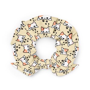 Può includere: Un elastico per capelli giallo con un motivo ripetuto di gatti bianchi con orecchie arancioni che spuntano da sopra e impronte di zampe nere.